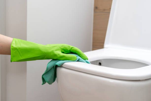 Close up view woman hand cleaning white ceramic toilet bowl at home. Cropped shot of female in green protective gloves using a blue rag for disinfecting the restroom seat
