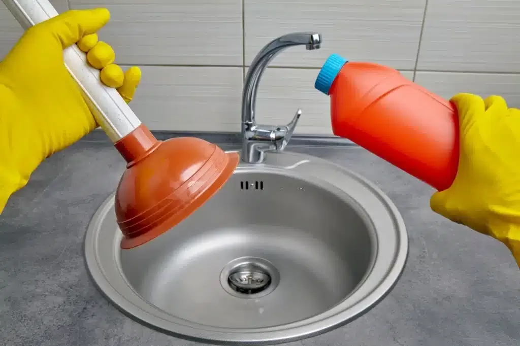 A plumber standing infront of a sink with a plunger and a orange bottle in his hand